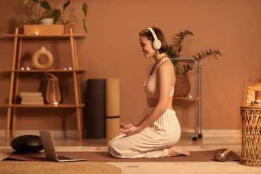 Akşamları evde kulaklık ve dizüstü bilgisayarla meditasyon yapan güzel genç bir kadın.
