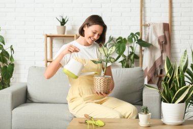 Kanepede oturan ve evdeki çiçekleri sulayan gülümseyen genç kadın.