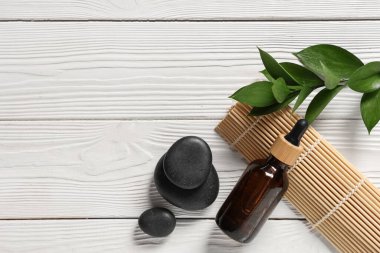 Beyaz ahşap arka planda bir şişe temel yağ, spa taşları, bambu hasırı ve bitki dalı.