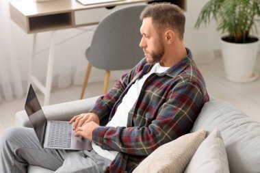 Yakışıklı sakallı adam oturma odasındaki gri koltukta dizüstü bilgisayar kullanıyor.