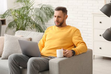 Elinde kahve olan genç adam oturma odasında dizüstü bilgisayar kullanıyor.