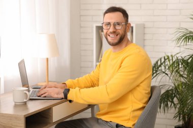 Genç adam iş yerinde oturup evde dizüstü bilgisayarla çalışıyor.