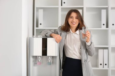 Ofisteki soğutucudan su içen gülümseyen genç kadın.