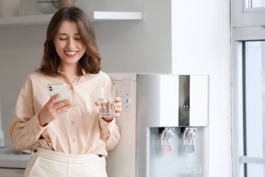 Soğutucudan su içen ve mutfakta cep telefonu kullanan genç bir kadın.