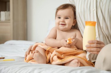 Annesi evde bir şişe krema tutarken bebek havluyla yatakta oturuyor. Yakın çekim.