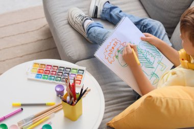 Küçük çocuk kanepede oturuyor ve oturma odasında çizim yapıyor.