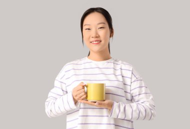 Genç Asyalı kadın gri arka planda sarı bardaktan kahve içiyor.