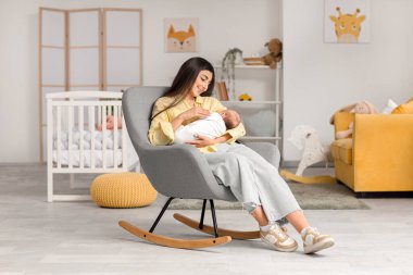 Kadın bebeğini emzirirken evdeki koltukta oturuyor.