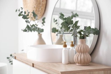 Lavabo, ayna ve yeşil dallı vazolar. Banyodaki beyaz duvarın yanındaki çekmecelerin yanında.