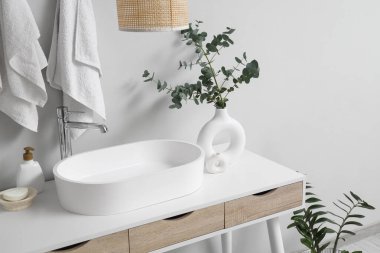 Lavabolu masa, kozmetik ürünleri şişesi ve banyoda beyaz duvarın yanında okaliptüs dalları olan vazo.