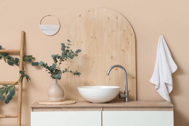 Banyodaki bej duvarın yanında yeşil dallı vazo ve lavabolu çekmeceler.