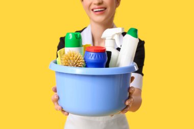 Sarı arka planda temizlik ürünleri ve aletlerle dolu bir leğen tutan güzel genç oda hizmetçisi.