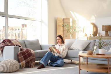 Güzel genç bir kadın, büyük pencereli şık bir oturma odasında yerde kitap okuyor.