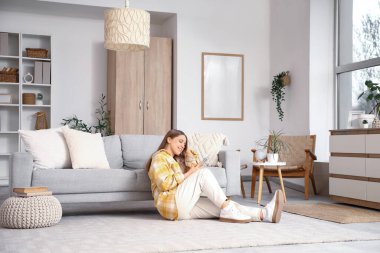 Güzel genç bir kadın büyük pencereli şık bir oturma odasında yerde cep telefonu kullanıyor.