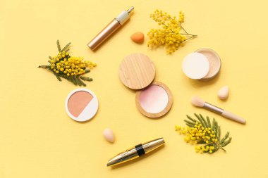 Sarı arka planda makyaj süngerleri ve mimoza çiçekleri olan dekoratif kozmetik ürünler.