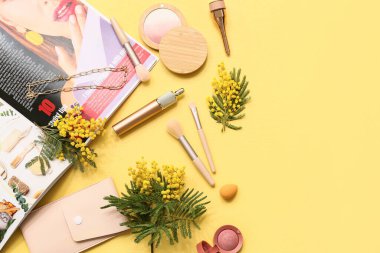 Sarı arka planda zincirli kolye, dergi ve mimoza çiçekleri olan dekoratif kozmetik ürünler.