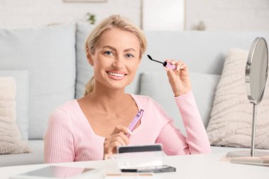 Güzel bir kadın oturma odasında aynaya bakarken maskara sürüyor.