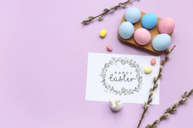 Wooden holder with Easter eggs, greeting card and pussy willow branches on lilac background