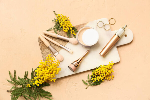 Доска с декоративной косметикой, стильными кольцами и цветами мимозы на бежевом фоне