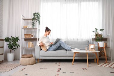 Evdeki kanepede dizüstü bilgisayarı olan güzel genç bir kadın.