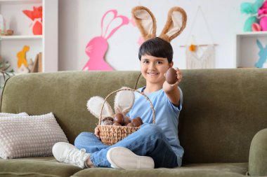 Oturma odasındaki kanepede oturan sepet dolusu çikolatalı tatlı yumurtalı tavşan kulaklı şirin çocuk. Paskalya kutlaması