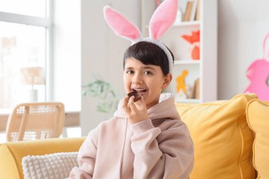 Tavşan kulaklı tatlı çocuk oturma odasında çikolatalı yumurta yiyor. Paskalya kutlaması