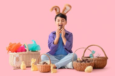 Tatlı çikolata yumurtası, paskalya sepetleri ve pembe arka planda oturan oyuncaklarla tavşan kulaklı tatlı çocuk.