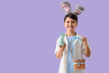 Tavşan kulaklı şirin çocuk sepet tutuyor, yanında çikolata yumurtaları ve leylak arka planda oyuncak tavşan var. Paskalya kutlaması