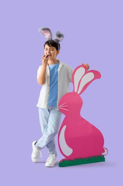 Tavşan kulaklı şirin çocuk, leylak arka planda kağıttan tavşanla çikolatalı yumurta yiyor. Paskalya kutlaması