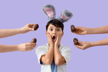 Leylak arkaplanlı çikolata yumurtalı tavşan kulaklı ve elleri olan şok olmuş bir çocuk. Paskalya kutlaması