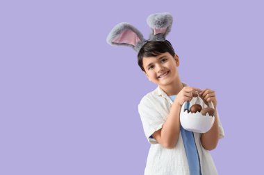 Tavşan kulaklı şirin çocuk, leylak arkaplanlı çikolata yumurtalı sepet tutuyor. Paskalya kutlaması