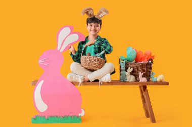Tavşan kulaklı, sepet sepetinde çikolatalı yumurta ve sarı arka planda duran kağıt tavşanlı sevimli çocuk. Paskalya kutlaması