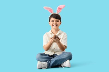 Mavi arka planda oturan, çikolata yumurtalı, tavşan kulaklı tatlı çocuk. Paskalya kutlaması
