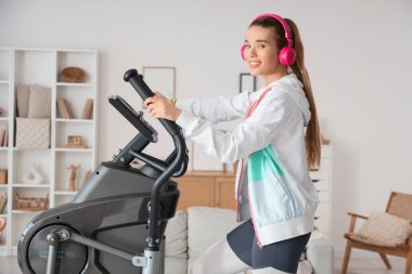 Evde kulaklıklı, modern egzersiz bisikleti eğitimi alan genç bir bayan.