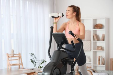 Spor yapan genç bir kadın modern egzersiz bisikletinde su içiyor.