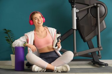 Su şişesi ve kulaklıklı genç bir bayan modern egzersiz bisikletinin yanında oturuyor.