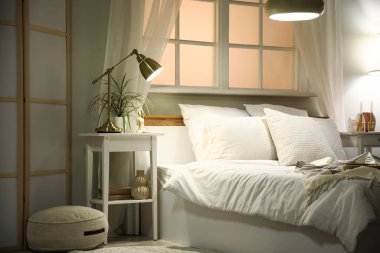 Komodininin yanında saksı, büyük yatağın yanında şilte ve akşam lambaları.