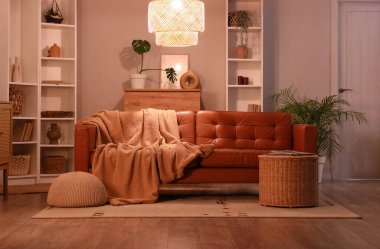 Akşamları kahverengi kanepe, hasır sepet, battaniye ve parlayan lambayla modern oturma odasının içinde.