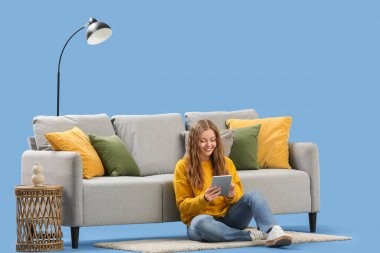 Tablet bilgisayarlı güzel genç bir kadın mavi arka planda şık gri kanepenin yanında oturuyor.