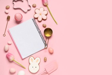 Tarifler, pişirme araçları, paskalya yumurtaları ve pembe arka planda kurabiyeler için boş defter içeren kompozisyon