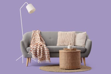 Yastıklı gri kanepe, hasır sepet ve mor arka planda yer lambası.