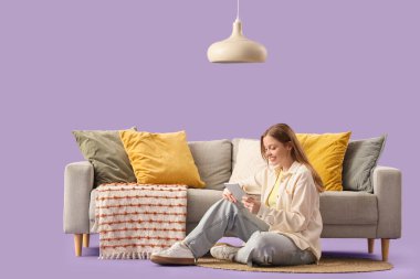 Genç kadın tablet bilgisayar kullanıyor ve mor arka planda gri kanepenin yanında yerde yatıyor.