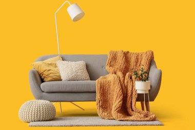 Sarı arka planda battaniyeli, yastıklı, çiçekli ve zemin lambalı rahat gri kanepe.