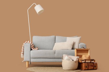 Kahverengi arka planda battaniyeli, sepetli, bavullu ve zemin lambalı rahat gri kanepe.