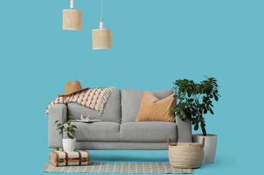 Mavi arka planda battaniyeli, valizli, sepetli ve çiçekli rahat gri kanepe.