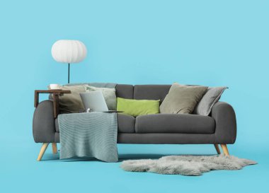 Mavi arka planda battaniyeli, yastıklı, laptoplu ve zemin lambalı rahat gri kanepe.