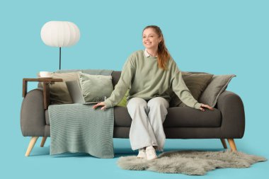 Gri kanepede oturan genç kadın battaniyeli ve mavi arka planda yastıklı.