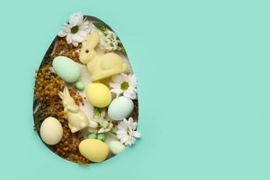 Paskalya yumurtaları, dekoratif tavşanlar ve çiçekler turkuaz arka planda yumurta şeklindeki delikten görülebilir.