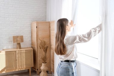 Evde perdeleri açan genç bir kadın.