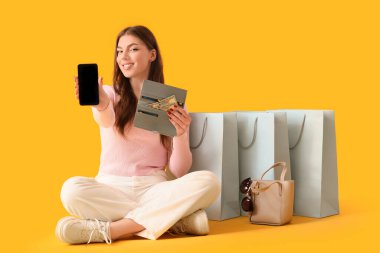 Çantalı ve cep telefonlu genç bir kadın sarı arka planda alışveriş yapıyor.